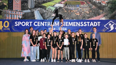 Gruppe junger Sportler mit Medaillen auf Bühne, während eine Akrobatin in der Luft balanciert. Hintergrund mit Sportzentrum.