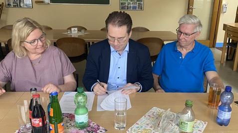 Drei Personen sitzen an einem Tisch mit Getränken und Unterlagen; eine Person unterschreibt ein Dokument.