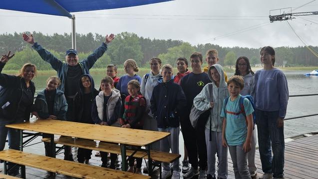 Gruppe von Jugendlichen und Erwachsenen unter einem blauen Sonnenschutz am Wasser, mit Wassersportgeräten im Hintergrund.