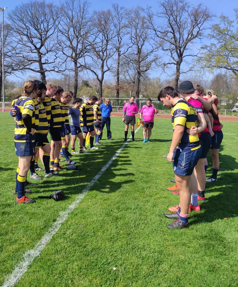 Zwei Rugby-Teams in gestreiften Trikots stehen auf einem Feld, während Schiedsrichter und Zuschauer beobachten.