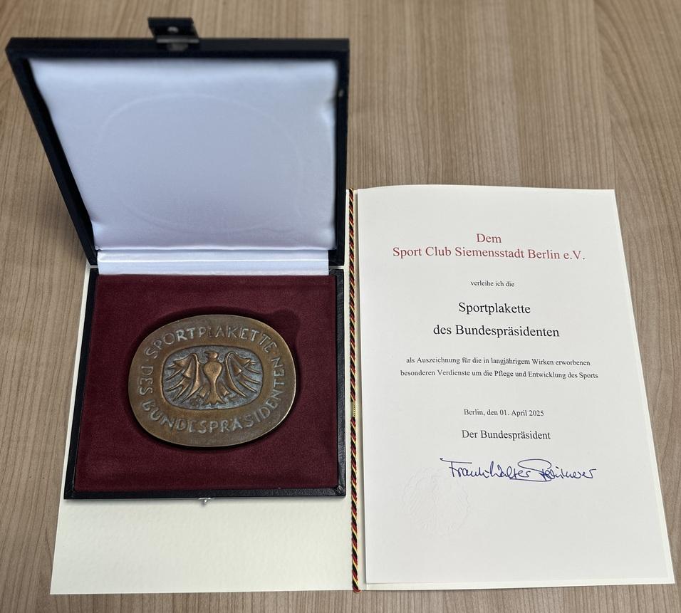 Medaille der Sportplakette des Bundespräsidenten in der Box, daneben ein Urkundendokument auf einem Tisch.