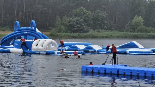 Gruppe von Personen auf einem schwimmenden Hindernisparkour im Wasser, umgeben von Bäumen und einem grauen Himmel.