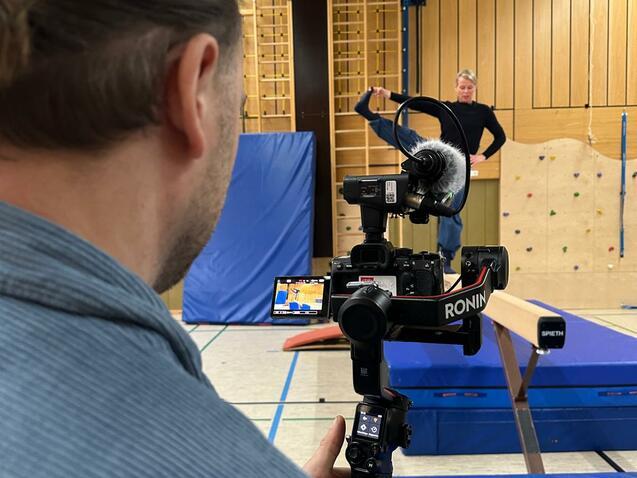 Person filmt eine Gymnastikübung in einer Turnhalle mit Kamera auf einem Stabilisator und einem Balancebalken im Vordergrund.