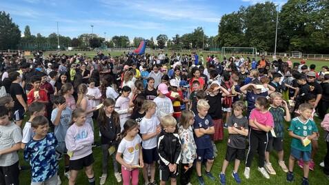 Große Gruppe von Kindern in sportlicher Kleidung, versammelt auf einem Rasensportplatz unter blauem Himmel.