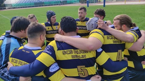 Rugby-Mannschaft in schwarz-gelben Trikots bildet einen Kreis auf dem Spielfeld und bespricht sich vor dem Spiel.