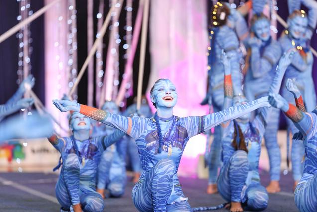 Tanzende Performer in blauen Kostümen mit Gesichtsbemalung, umgeben von festlicher Dekoration und Lichtern auf der Bühne.