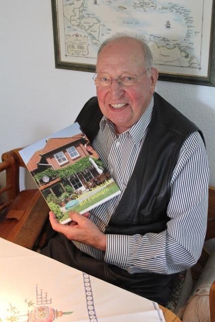 Lächelnder älterer Mann hält ein Buch mit einem Hauscover in der Hand, sitzt an einem Tisch mit bestickter Tischdecke.
