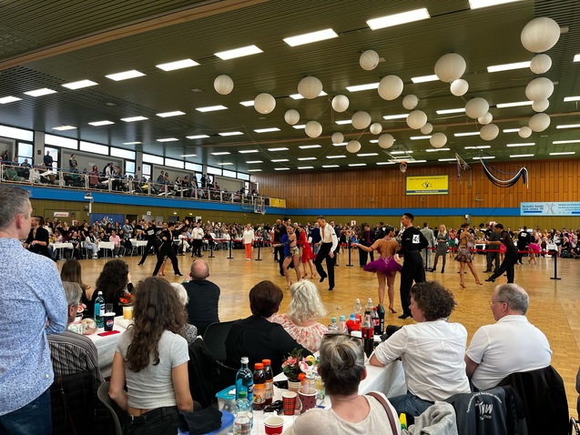 Tanzpaare führen synchron Tänze auf einem Wettbewerb auf, Zuschauer betrachten das Geschehen in einer großen Halle.