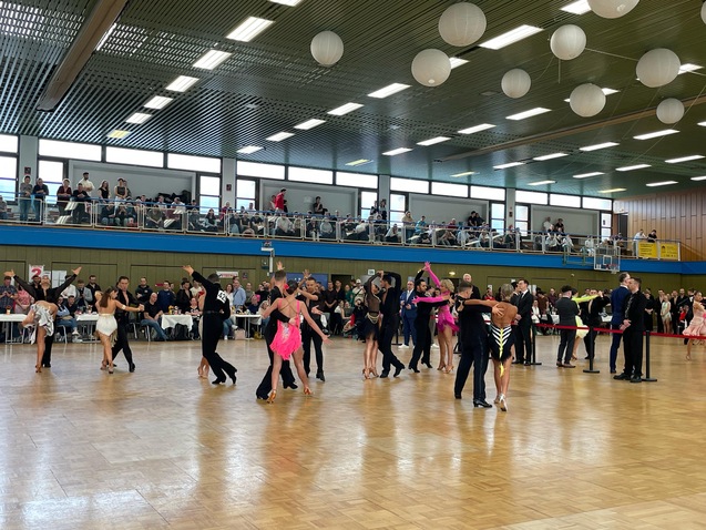 Tanzpaare in verschiedenen Outfits zeigen einen Wettbewerb in einem großen Saal mit Zuschauern auf der Tribüne.
