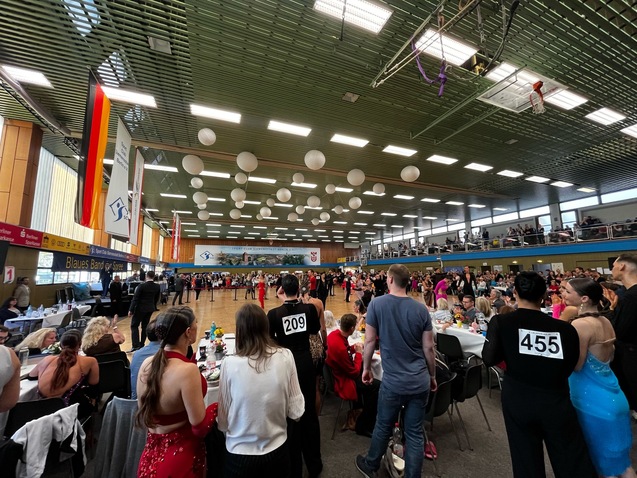 Tanzwettbewerb in einer großen Halle mit Zuschauern, Tänzern und festlich eingedeckten Tischen im Hintergrund.