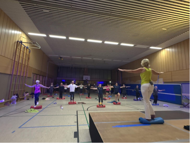 Gruppentraining in einer Sporthalle: Teilnehmer stehen auf Balance-Kissen und folgen einer Trainerin mit erhobenen Armen.