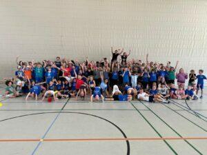 Gruppenfoto von über 70 Kindern in blauen T-Shirts, die freudig in einer Sporthalle posieren.