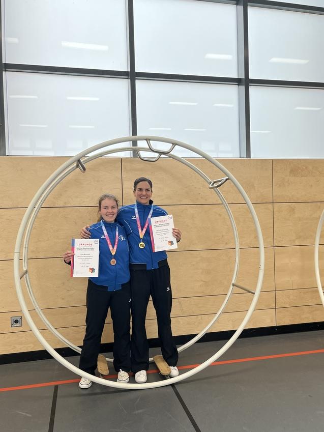 Zwei Athletinnen in blauen Sportjacken stehen in einem großen Holzring und halten Medaillen und Urkunden in den Händen.