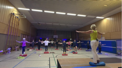 Gruppentraining in einer Sporthalle: Teilnehmer stehen auf Balance-Kissen und folgen einer Trainerin mit erhobenen Armen.