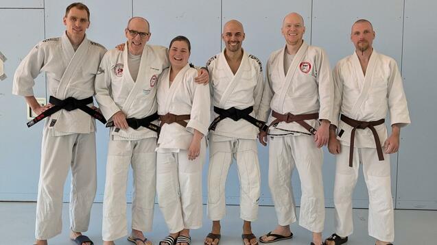 Gruppe von fünf Judokas in Judogis, zwei mit braunen und drei mit schwarzen Gürteln, stehen lächelnd in einer Trainingshalle.