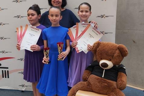 Drei M&auml;dchen in Tanzkleidern halten Pokale und Urkunden, stehen mit einer Frau vor einem Banner des Tanzsportverbands.