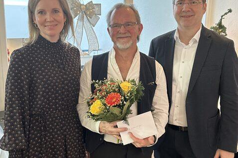 Drei Personen stehen in einem festlichen Raum, einer h&auml;lt einen Blumenstrau&szlig; und eine Karte. Weihnachtsdekoration im Hintergrund.