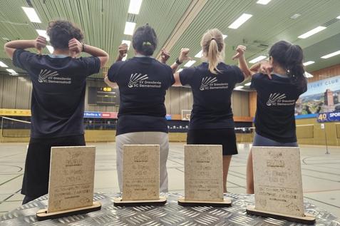 Vier Personen in T-Shirts mit Vereinslogo zeigen ihre Medaillen in einer Sporthalle. Auf einem Tisch stehen drei Pokale.