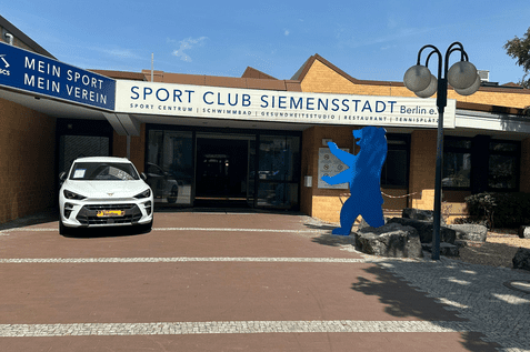 Eingangsbereich des Sport Club Siemensstadt in Berlin mit einem weißen Auto und einer blauen Bärenfigur.
