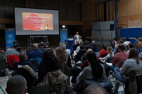 Vortrag zu Peer-Gewalt mit Projektorbildschirm und Publikum in einem Sportraum. Der Redner steht hinter einem Tisch.