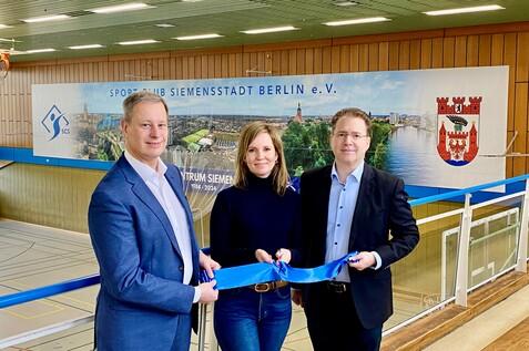 Drei Personen halten ein blaues Band in einer Sporthalle, im Hintergrund ein Banner des Sport Clubs Siemensstadt Berlin.