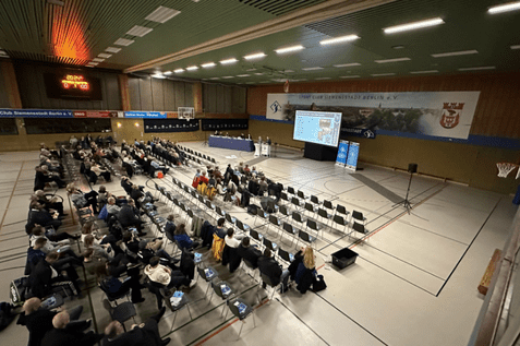 Versammlung in einer Sporthalle mit zahlreichen St&uuml;hlen und einer B&uuml;hne im Hintergrund. Zuschauer folgen der Pr&auml;sentation.