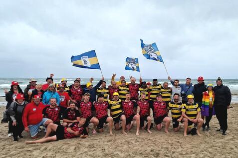 Gruppenantwortung von Rugbyspielern in Teamtrikots mit Fahnen am Strand, umgeben von Unterst&uuml;tzern in bunten H&uuml;ten.