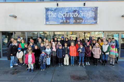 Gruppe von Kindern und Erwachsenen steht vor einem Geb&auml;ude mit Plakat f&uuml;r &bdquo;Die Schneek&ouml;nigin&ldquo;, ein Tanzprojekt.