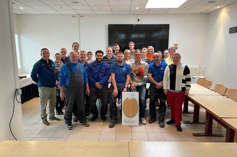 Gruppenfoto von 25 Personen in einem Raum, einige halten Blumen, andere stehen um ein Schild in der Mitte.