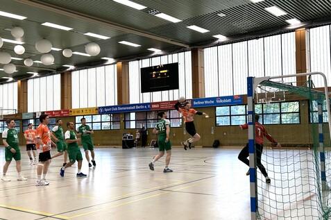 Aktion im Handballspiel: Spieler in orange springt zum Torwurf, w&auml;hrend gr&uuml;ne Spieler und Torwart beobachten.