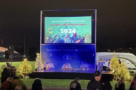 B&uuml;hne mit einem digitalen Banner f&uuml;r das Spandauer Weihnachtssingen 2024, umgeben von beleuchteten Tannenb&auml;umen.