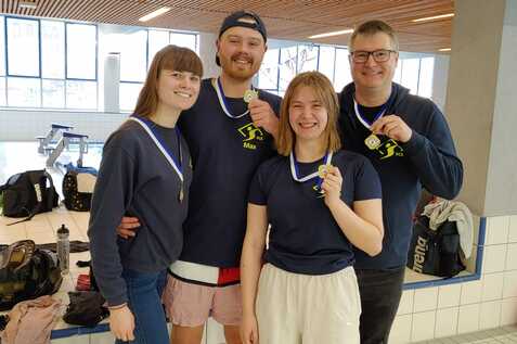 Vier Personen in Sportkleidung halten Medaillen in einer Schwimmhalle, Hintergrund zeigt Schwimmbecken und Taschen.