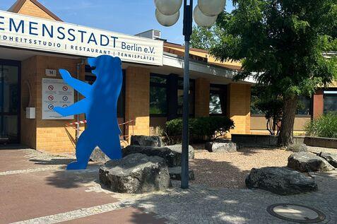 Blaue Figur vor einem Gebäude mit dem Schriftzug „E-SPORTS STADT Berlin e.V.“ und Laternen im Freien.