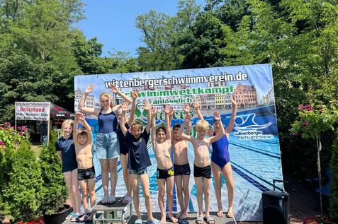 Gruppe von Kindern in Badebekleidung auf einem Siegerpodest, jubelnd vor einem Banner des Wittenberger Schwimmvereins.