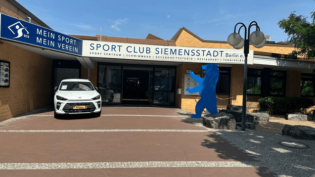 Eingangsbereich des Sport Club Siemensstadt in Berlin mit einem weißen Auto und einer blauen Bärenfigur.