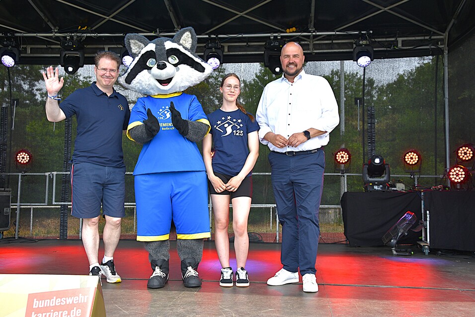 Drei Personen stehen auf einer Bühne mit Lichttechnik, ein Maskottchen in blauer Kleidung winkt.