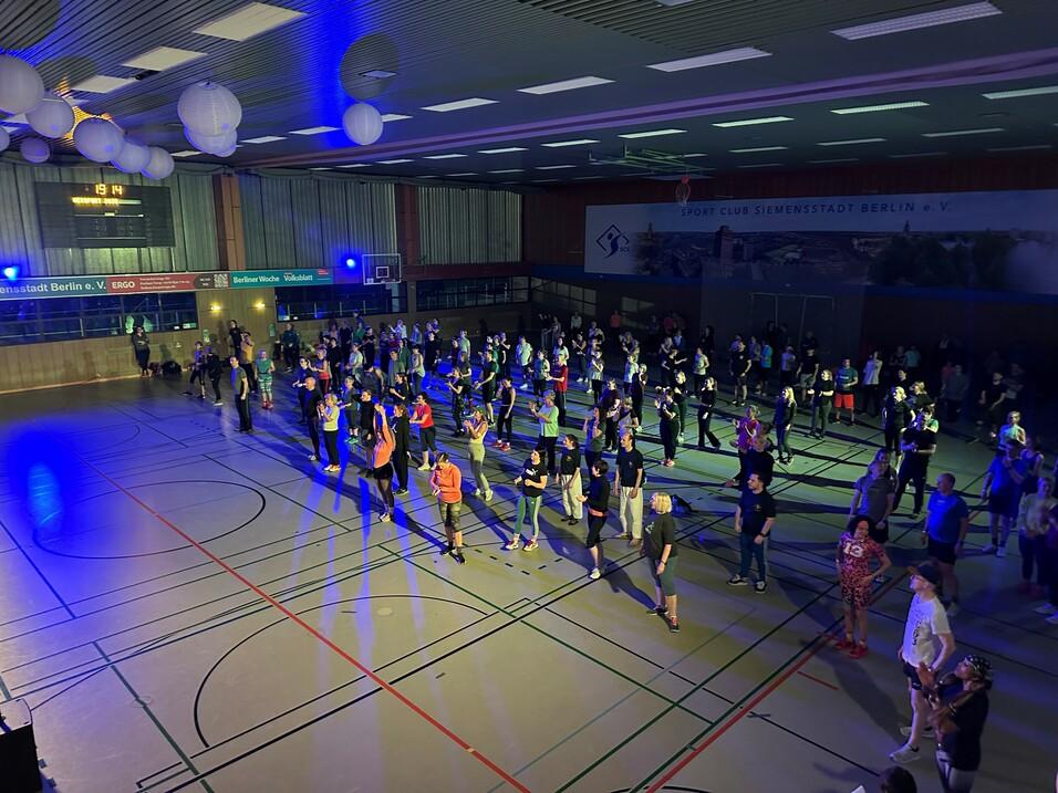 Menschen tanzen in einem hell beleuchteten Sporthall mit bunten Lichtern und dekorativen Laternen.