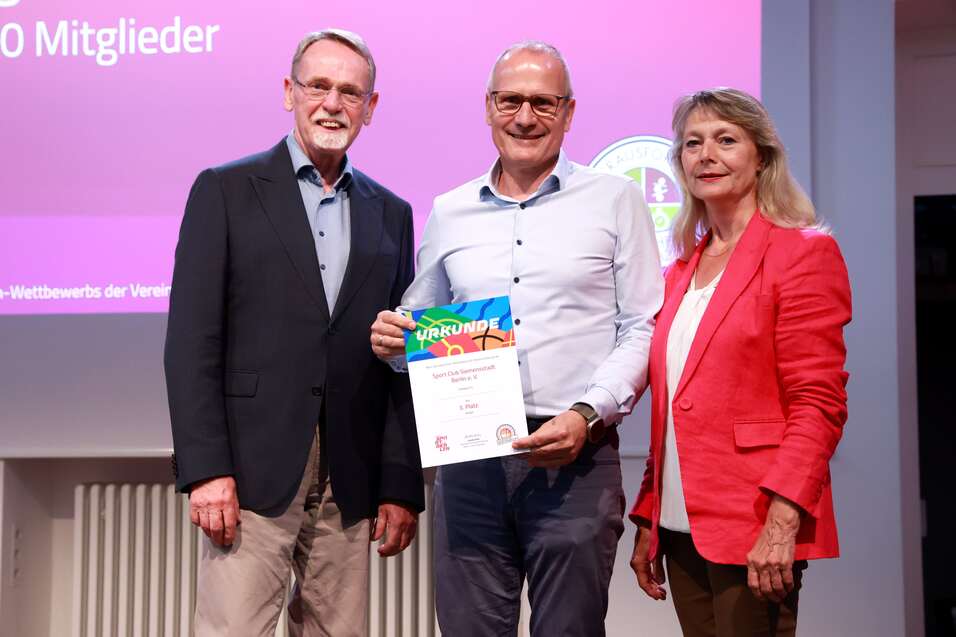 Drei Erwachsene stehen zusammen auf einer Bühne und halten eine Urkunde des Vereinswettbewerbs in der Hand.