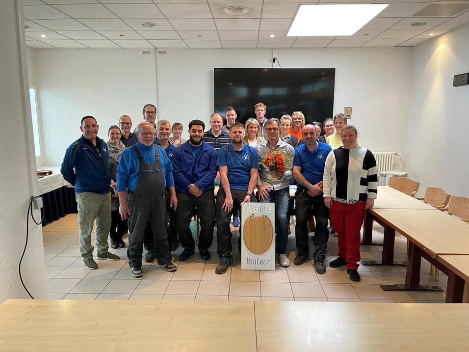 Gruppenfoto von 25 Personen in einem Raum, einige halten Blumen, andere stehen um ein Schild in der Mitte.