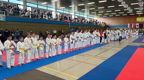 Gruppe von jungen Karatekas in weißen Gi mit verschiedenen Gürtelfarben in einer Sporthalle bei einer Veranstaltung.