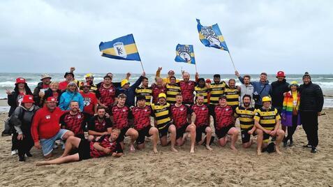 Gruppenantwortung von Rugbyspielern in Teamtrikots mit Fahnen am Strand, umgeben von Unterstützern in bunten Hüten.