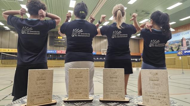 Vier Personen in T-Shirts mit Vereinslogo zeigen ihre Medaillen in einer Sporthalle. Auf einem Tisch stehen drei Pokale.