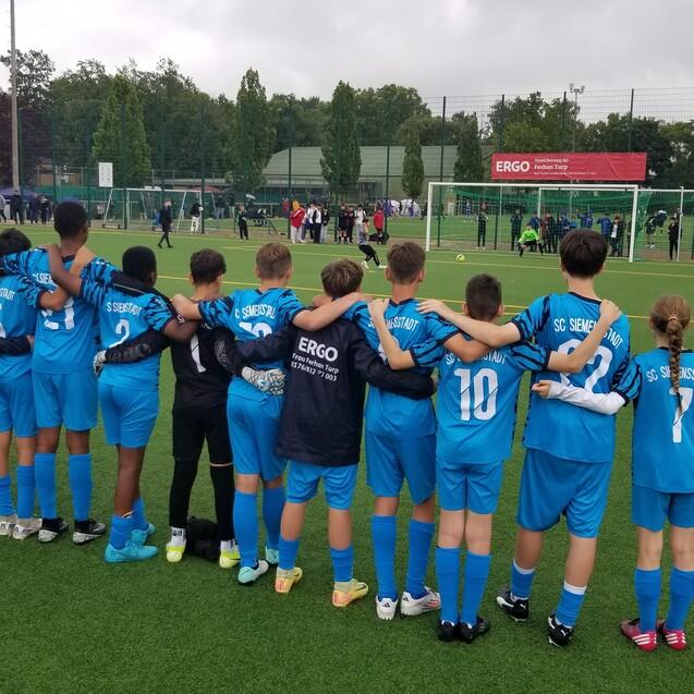 Gruppe von Kindern in blauen Fu&szlig;balltrikots, die Arm in Arm am Spielfeldrand stehen und ein Spiel beobachten.