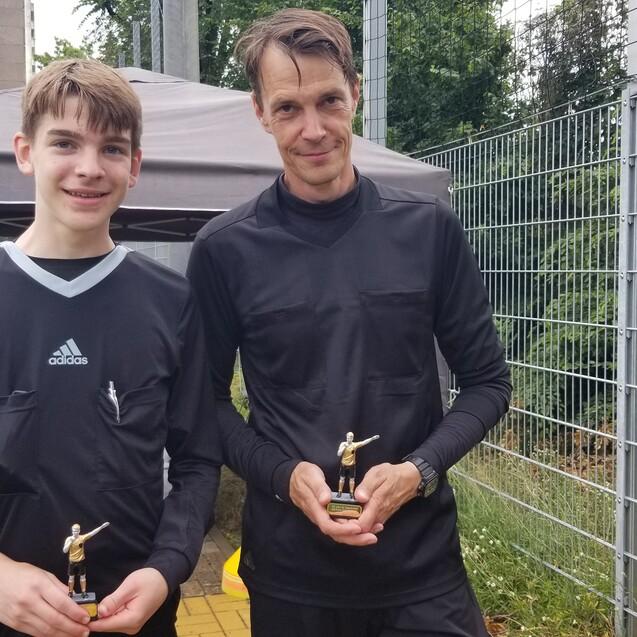 Zwei Fu&szlig;ballspieler in schwarzen Trikots halten Pokale in einer Au&szlig;enumgebung mit Sportplatz und Zelt im Hintergrund.