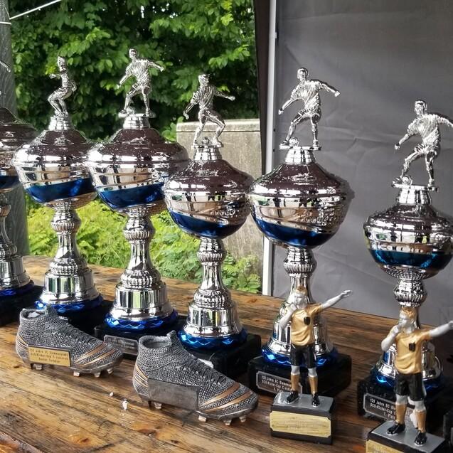 Reihe von silbernen Pokalen mit Figuren und einem Paar Fu&szlig;ballschuhen auf einem Holztisch im Freien.