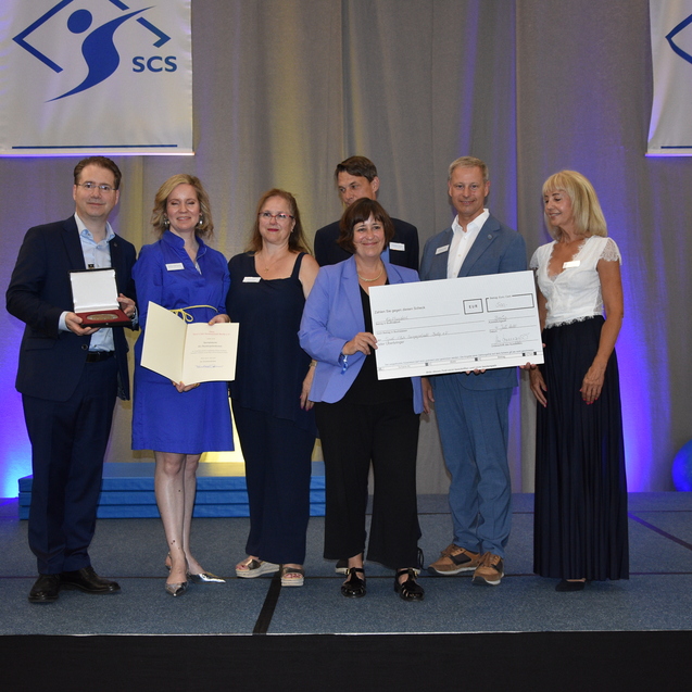 Gruppennahaufnahme von sechs Personen auf einer B&uuml;hne, die Auszeichnungen und einen Scheck f&uuml;r ein Projekt pr&auml;sentieren.