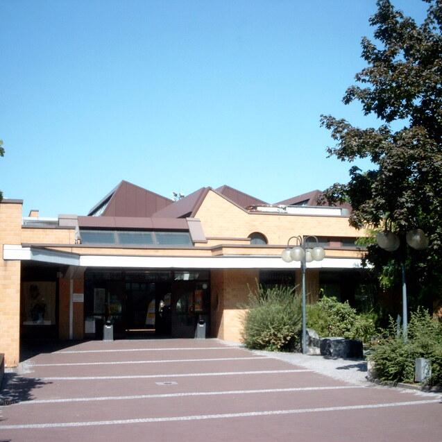 Eingangsbereich eines modernen Geb&auml;udes mit Glasfront, begr&uuml;ntem Umfeld und blauem Himmel.