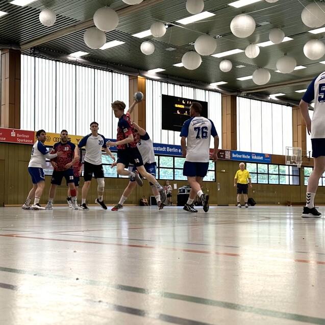 Handballspiel in einer Halle mit Spielern in wei&szlig;en und blauen Trikots, w&auml;hrend der Ball von einem Spieler geworfen wird.
