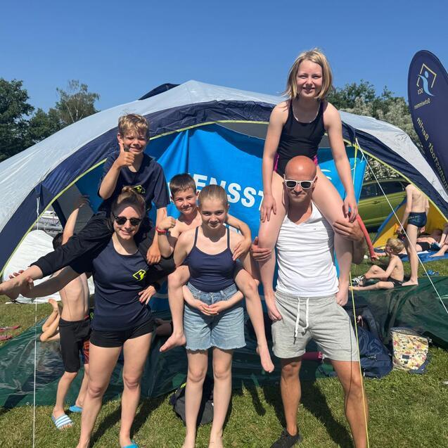 Gruppe von sechs Personen posiert unter einem Zelt bei sonnigem Wetter in einem Freibad.
