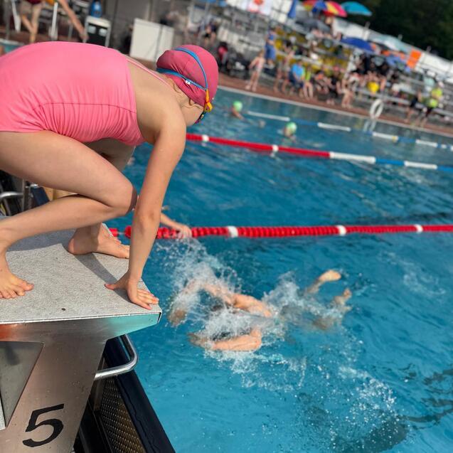 Ein M&auml;dchen in einem pinken Badeanzug springt vom Startblock ins Schwimmbecken, w&auml;hrend andere schwimmen.